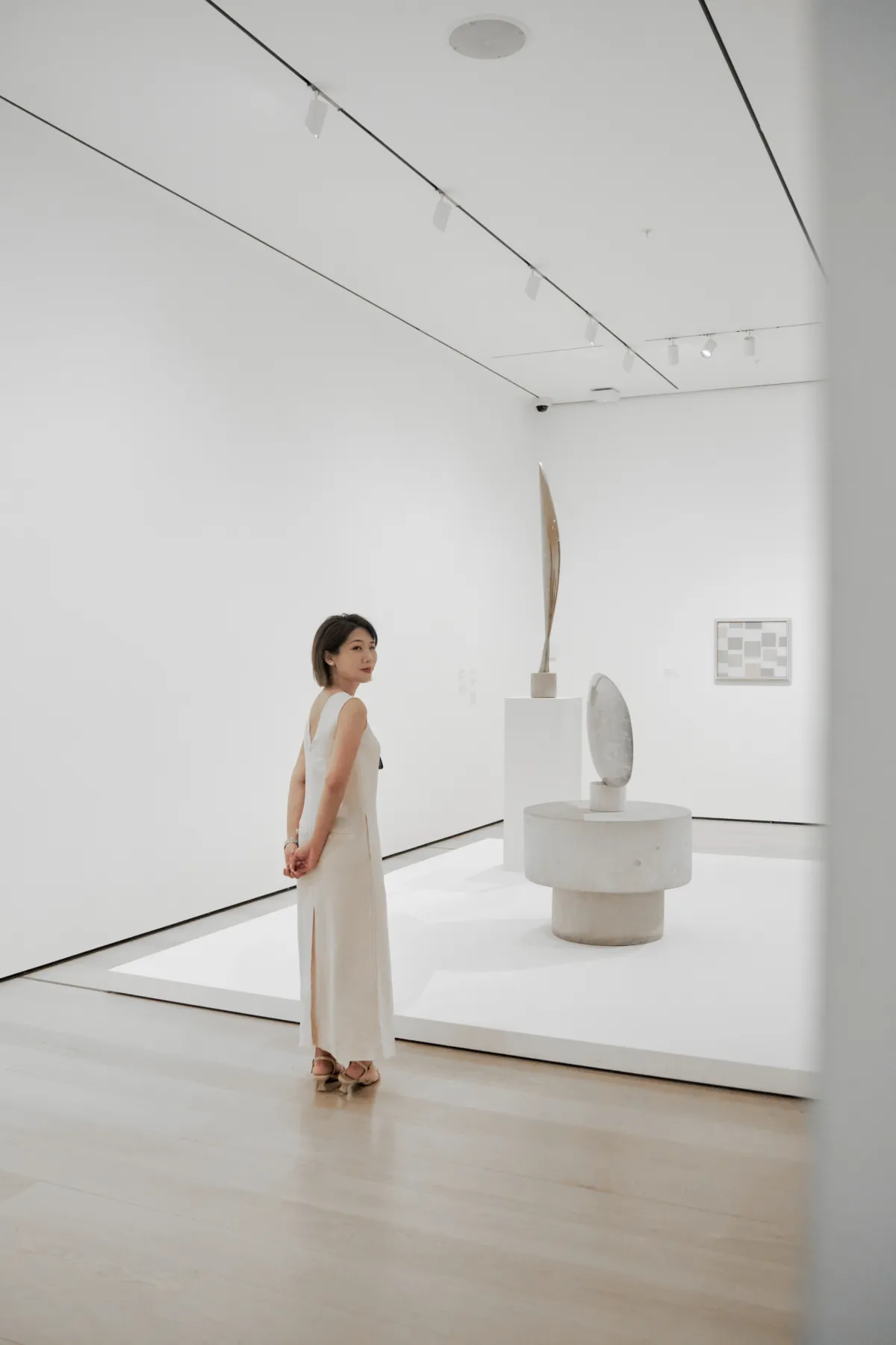 Janus in a long white dress, looking back over one shoulder in a white gallery with two sculptures on a plinth